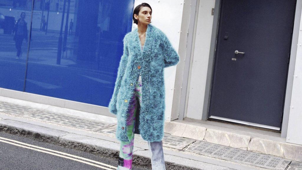 a woman standing on the streets in a long, fuzzy, blue cardigan and tie-dye pants
